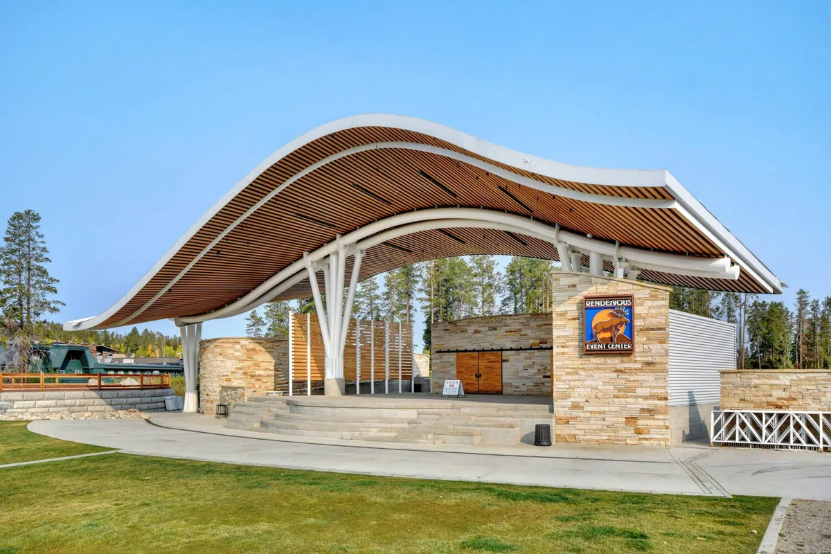 An exterior scene featuring a striking curved roof structure, a natural stone wall, and a stage area set outdoors. The setting suggests a vibrant community hub, ideal for concerts, festivals, and local events. The open air and scenic backdrop hint at the lively gathering opportunities available year-round in Winter Park's downtown and resort areas.