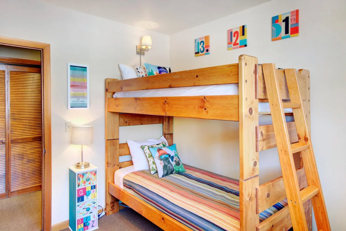 A playful twin bedroom setup with a bunk bed, colorful pillows, vibrant artwork on the wall, and a bedside lamp, creating an inviting space for children or friends. The décor is playful and cozy, perfect for storytime or relaxing after busy days.