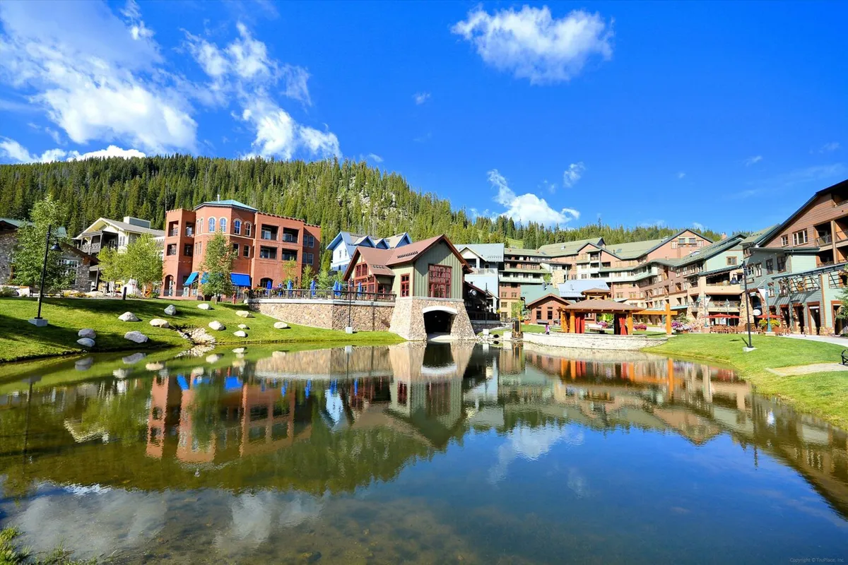 Colorful resort buildings reflect in the calm water of a landscaped pond, set against a backdrop of rolling mountains and lush greenery. People stroll along the water and explore the lively, cheerful village atmosphere.