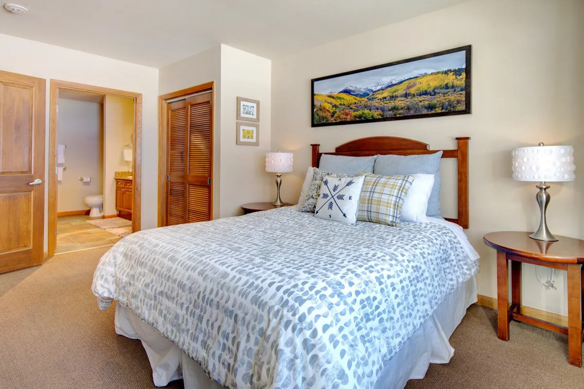 A tranquil bedroom featuring a bed with patterned bedding, soft landscape artwork above the headboard, and a nightstand with a warm-lit lamp. The room exudes comfort with plush linens and cozy mountain-inspired décor, ideal for restful sleep after a day of adventure.