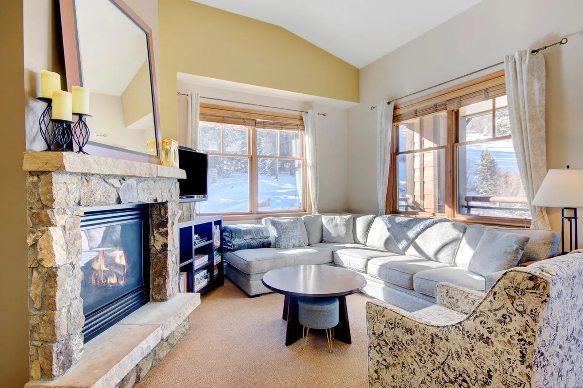 The image showcases a cozy, upscale living room with a large sectional sofa centered around a crackling fireplace. Expansive windows frame breathtaking mountain views, with snow gently falling outside. The inviting space is perfect for relaxing with loved ones after a day on the slopes, offering warmth and comfort in a picturesque alpine setting.