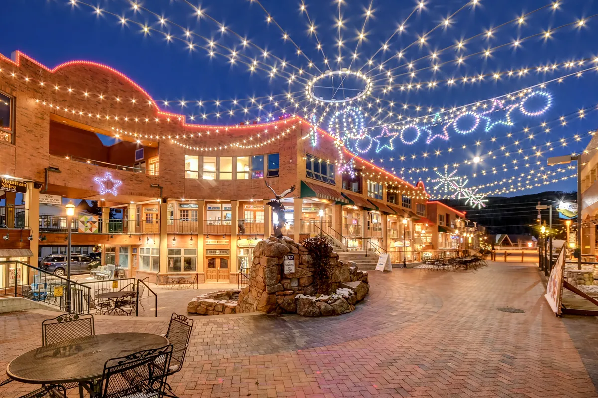 An inviting exterior scene of the resort village, with illuminated string lights, decorative storefronts, and a central fountain. The atmosphere is festive and social, suggesting a vibrant community space where families can gather and explore.