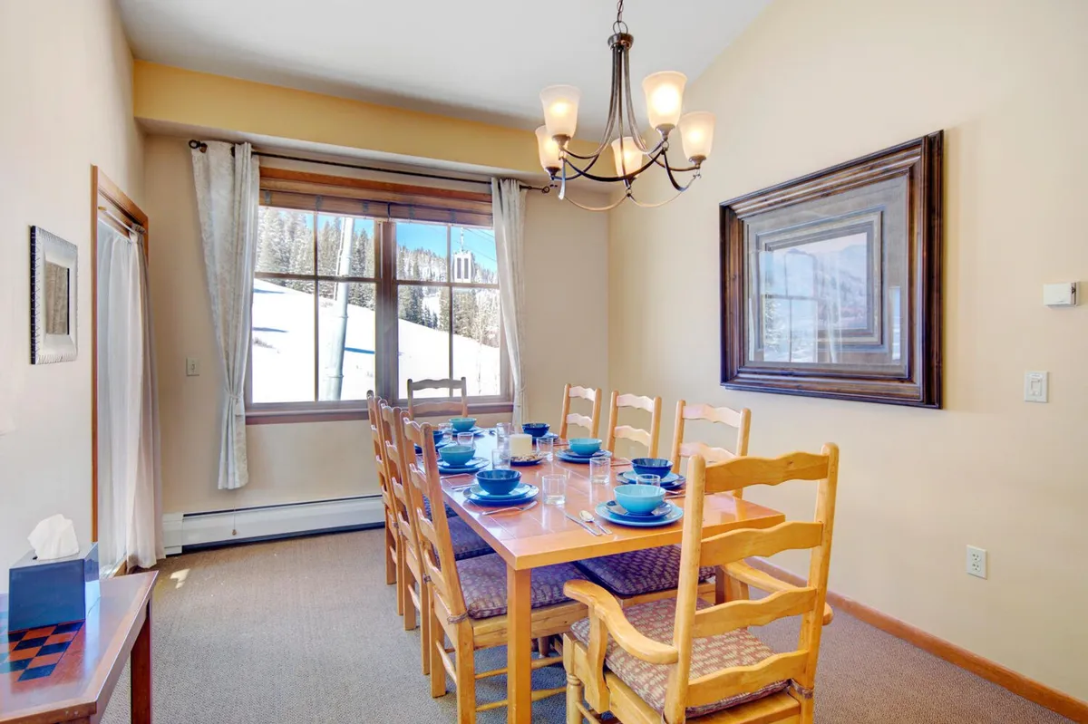A warmly lit dining room interior with a large table beneath a chandelier and a window framing scenic views. The scene evokes a sense of togetherness, ideal for group meals, celebrations, or relaxed evenings sharing stories.