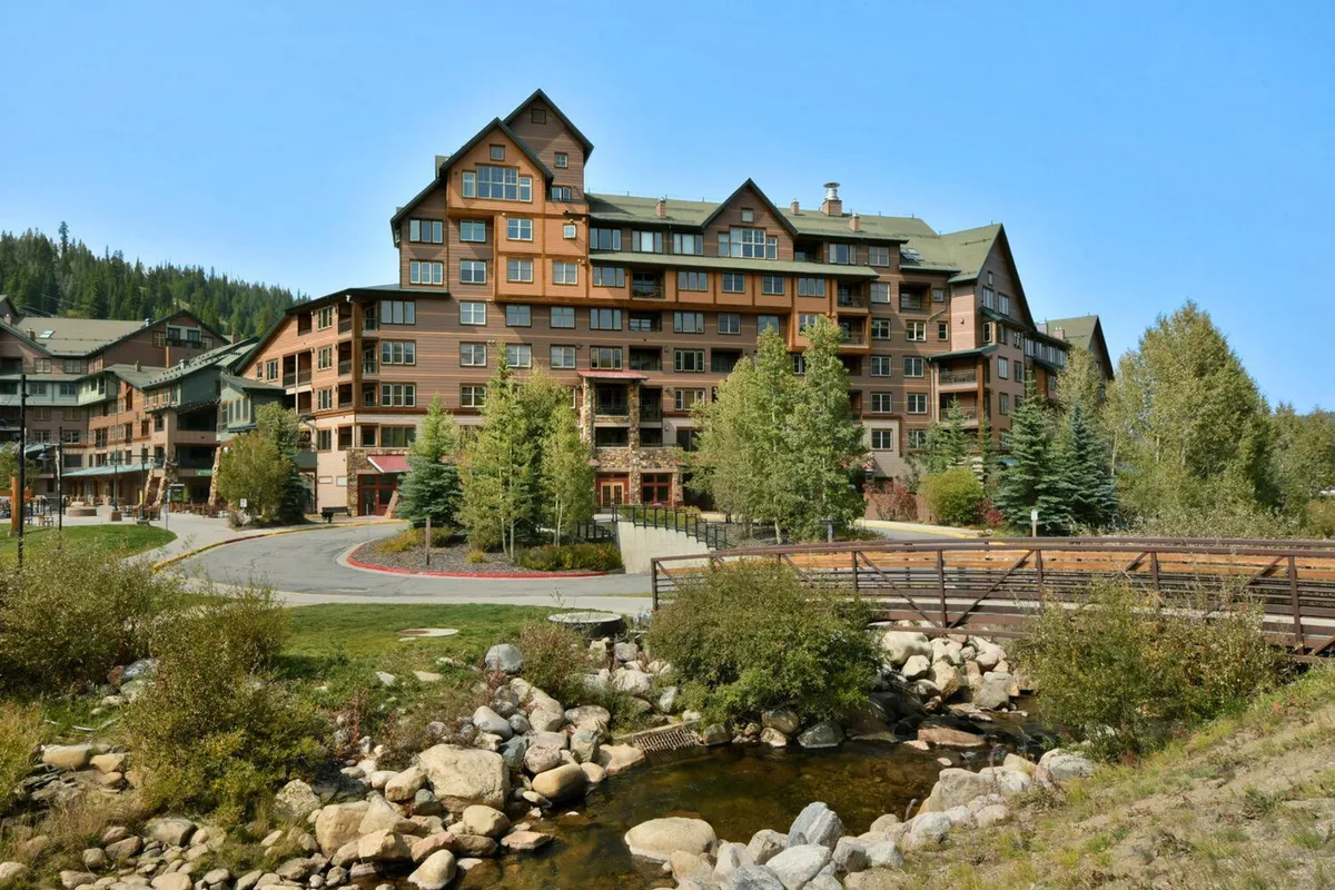 This exterior image features a multistory lodge nestled among trees, with a stream and a charming bridge in the foreground. The setting is tranquil, with the glow of a sunset casting warm light on the lodge, hinting at the beauty and serenity of the surrounding landscape. Seasonal activities like hiking and fishing are subtly suggested by the natural features.