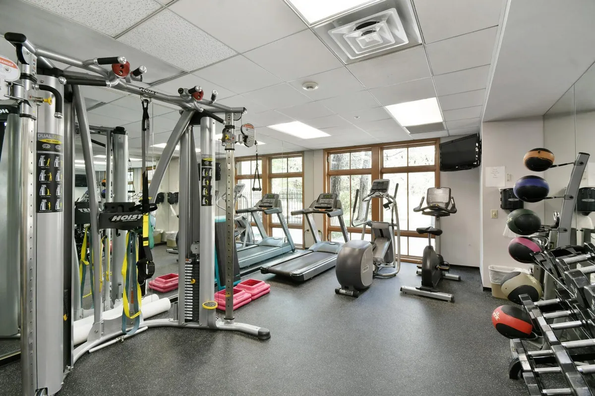 A vibrant and modern gym interior, featuring a variety of workout equipment such as treadmills and weight racks. Large windows bring in natural light and offer partial views of the outdoors, while a few guests can be seen enjoying their fitness routines.